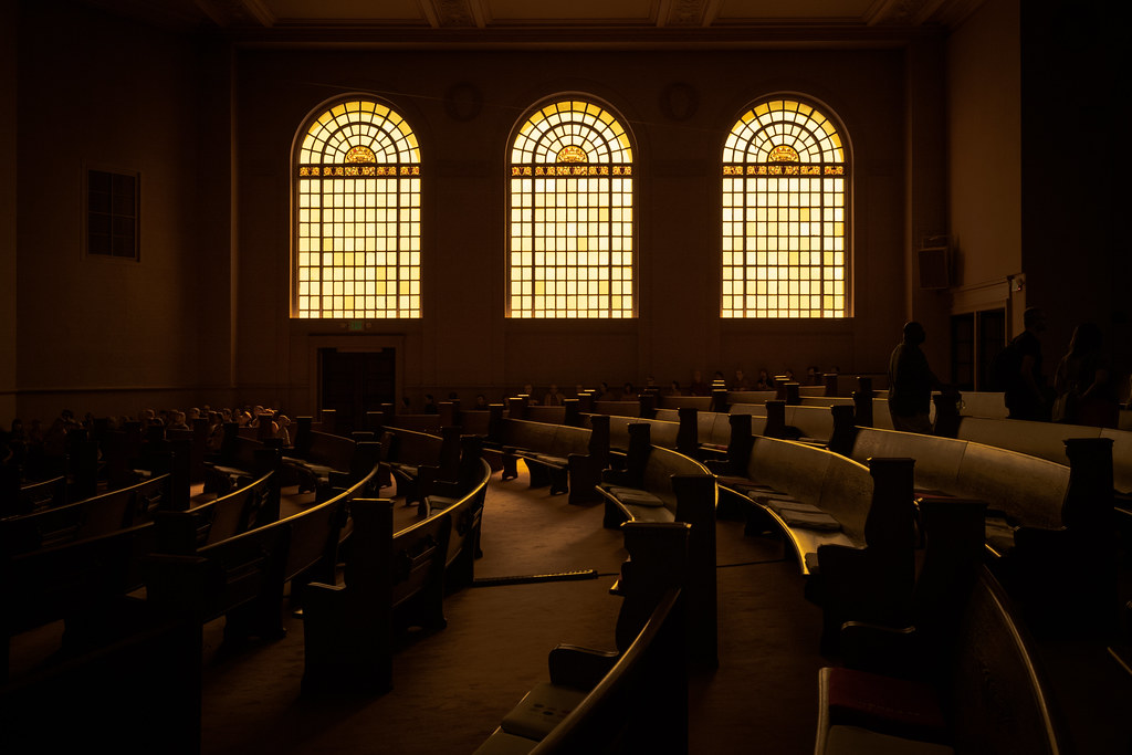 A photograph taken in the main room of the internet archive. backlit through 3 big windows, one can see the pews in the foreground
